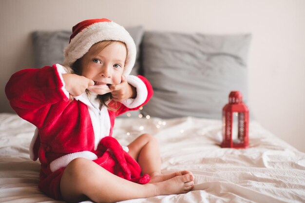 Niña linda usa sombrero rojo de santa claus y bata de baño sentada en la cama con decoración navideña