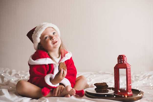 Niña linda usa sombrero rojo de santa claus y bata de baño sentada en la cama con decoración navideña