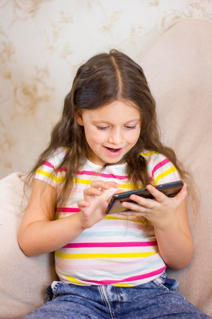 Foto una niña linda está usando un teléfono inteligente sentado en el sofá de casa y sonriendo