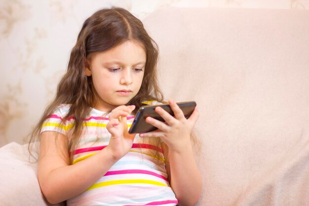 Foto una niña linda está usando un teléfono inteligente sentado en el sofá de casa y sonriendo