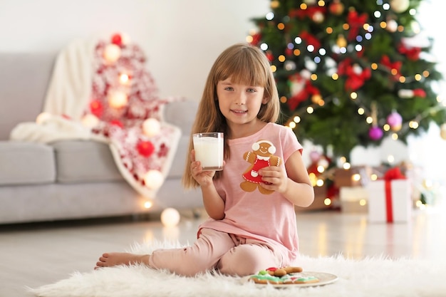 Niña linda con vaso de leche y galletas en la habitación decorada para Navidad