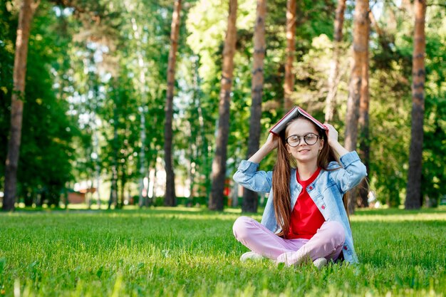 Foto niña linda en vasos con un libro sobre su cabeza en el parque de verano