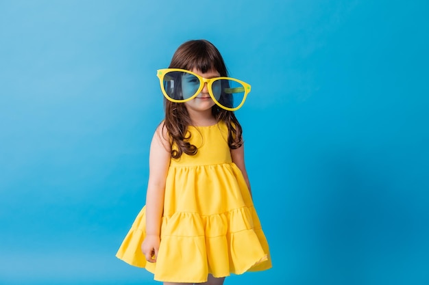 Niña linda con un vestido amarillo y enormes gafas de sol se encuentra en una pancarta de tarjeta de fondo azul