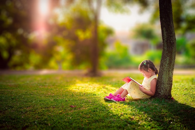 Una niña linda en un vestido amarillo leyendo un libro sentado bajo el árbol