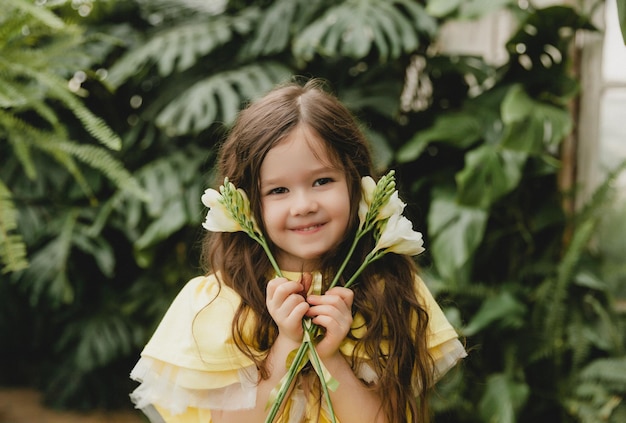 Niña linda con un vestido amarillo sosteniendo flores de primavera en sus manos de pie contra el fondo de las hojas