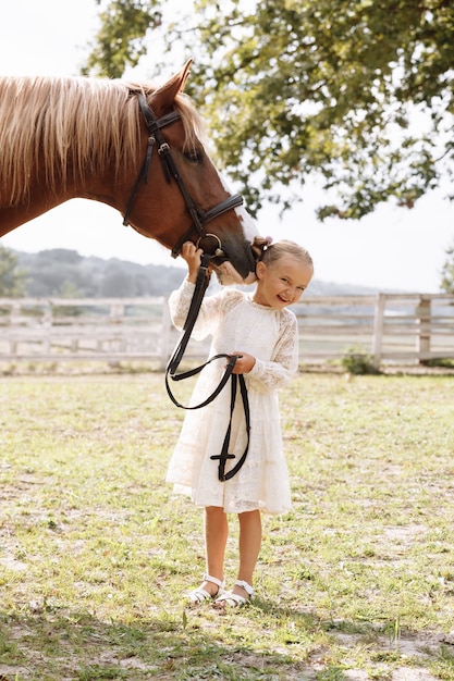 Foto niña linda en vestido blanco acariciando un caballo en el campo en la granja en un hermoso día de verano.