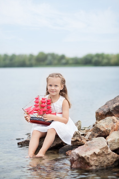 Niña linda en un vestido blanco y velas rojas. Un niño se sienta en una roca junto al mar con un barco de juguete.