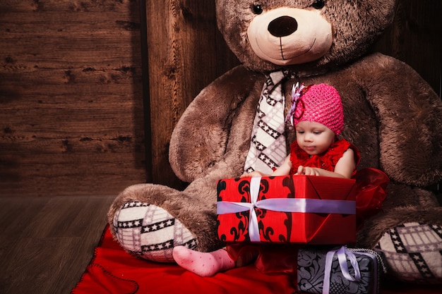 Niña linda en vestido rojo con gran oso de peluche y regalos