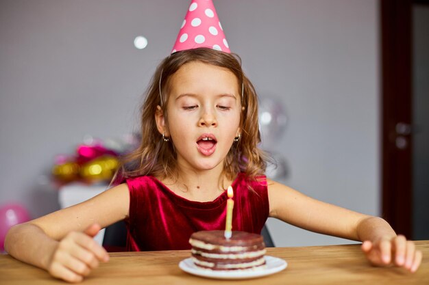 Una niña lleva un sombrero de cumpleaños mirando un pastel de cumpleaños y sopla velas para un concepto de fiesta de celebración