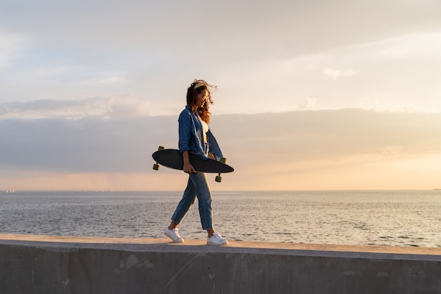 Niña llevar longboard al atardecer mirando al mar elegante skater femenino disfrutar de caminar junto al mar