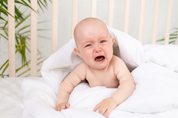 Una niña llorando en una manta en una cuna sobre una cama de algodón blanco de seis meses se ha despertado y está llamando a su madre