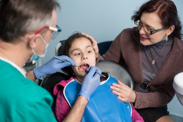 Niña con madre en la primera visita dental. Dentista masculino senior haciendo el primer chequeo para el paciente en el consultorio dental