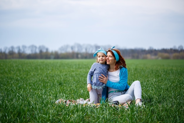 La niña y la madre se sientan en la colcha, la hierba verde en el campo, el clima soleado de primavera, la sonrisa y la alegría del niño, el cielo azul con nubes