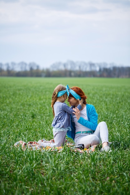 La niña y la madre se sientan en la colcha, la hierba verde en el campo, el clima soleado de primavera, la sonrisa y la alegría del niño, el cielo azul con nubes