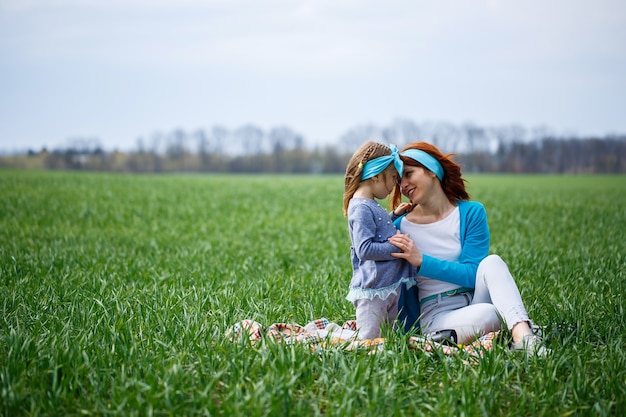 La niña y la madre se sientan en la colcha, la hierba verde en el campo, el clima soleado de primavera, la sonrisa y la alegría del niño, el cielo azul con nubes