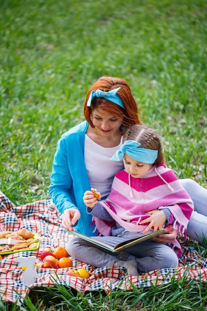 La niña y la madre se sientan en la colcha y leen un libro con un cuento de hadas, hierba verde en el campo, clima primaveral soleado, sonrisa y alegría del niño, cielo azul con nubes