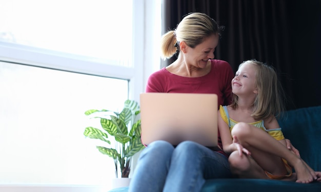La niña y la mamá están sentadas en el sofá con el portátil en el regazo y hablando