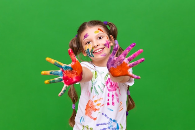Una niña manchada con pintura multicolor tira de sus manos hacia adelante y sonríe sobre un fondo verde aislado Creatividad artística infantil