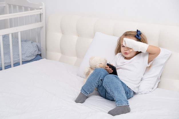 Niña con mano enyesada sentada en la cama con smartphone.