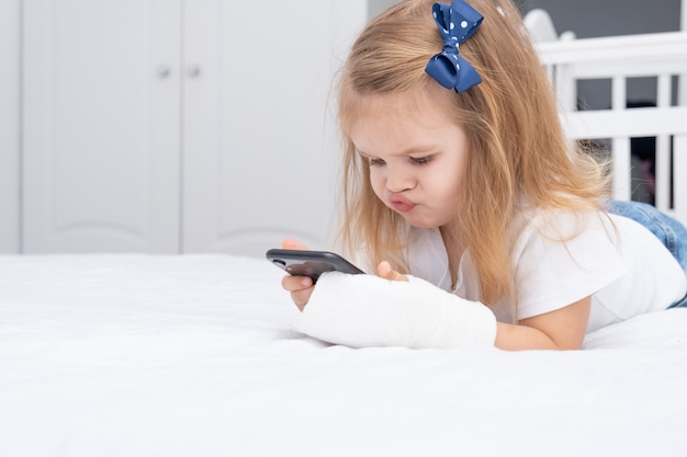 Niña con mano en yeso tendido en la cama con teléfono inteligente, viendo dibujos animados o videos educativos.