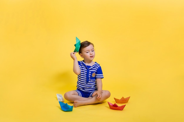 Una niña marinero con un bote de papel en sus manos está sentada en un espacio amarillo. Una hermosa chica en traje de marinero mira hacia otro lado. Origami de papel.