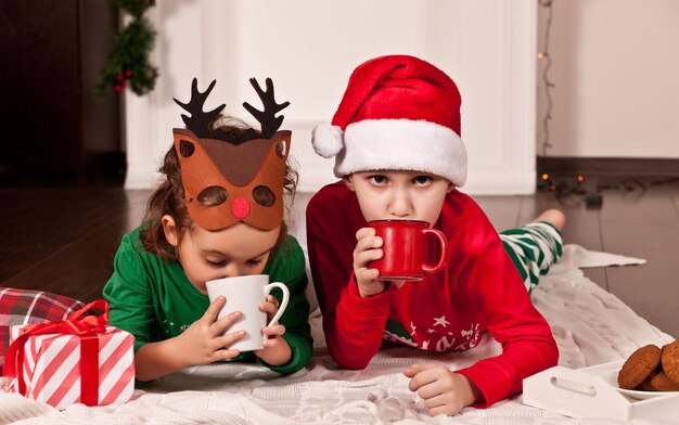 Niña con máscara de ciervo divertida y niño con gorro de Papá Noel y pijama de Navidad bebiendo cacao y comiendo galletas en casa