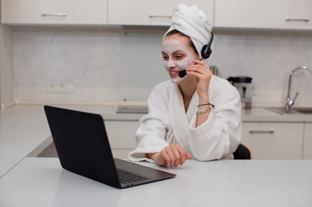 Una niña con una máscara hidratante en el rostro con una túnica blanca hablando de los auriculares trabajando detrás de una computadora portátil sentada en la cocina foto de alta calidad