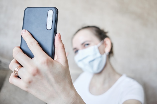 Una niña con una máscara médica está en cuarentena en su casa y se toma una selfie en un teléfono inteligente.