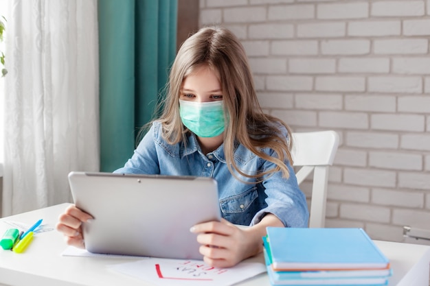 Una niña con una máscara médica está estudiando en casa con una tableta digital portátil y haciendo los deberes. Educación a distancia en línea