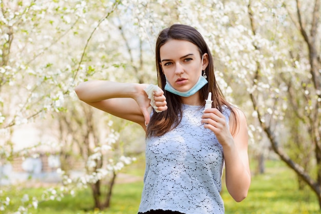 Niña en máscara médica mostrando los pulgares hacia abajo de pie en un jardín floreciente y sosteniendo spray para correr la nariz