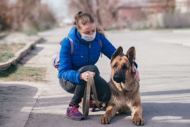 Una niña con una máscara médica protectora pasea a un perro en la calle. Ocio con una mascota durante la cuarentena. Modo de autoaislamiento y protección.