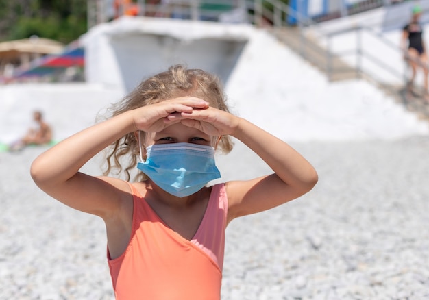 Niña en una máscara médica protectora en una playa de guijarros