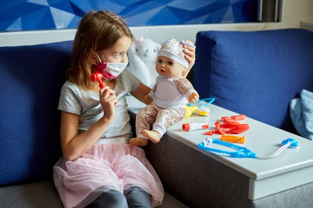 Una niña con máscara médica se sienta en el sofá, interpreta a un médico, trata y escucha a la muñeca con un estetoscopio en una máscara médica, en casa durante la cuarentena y la pandemia.