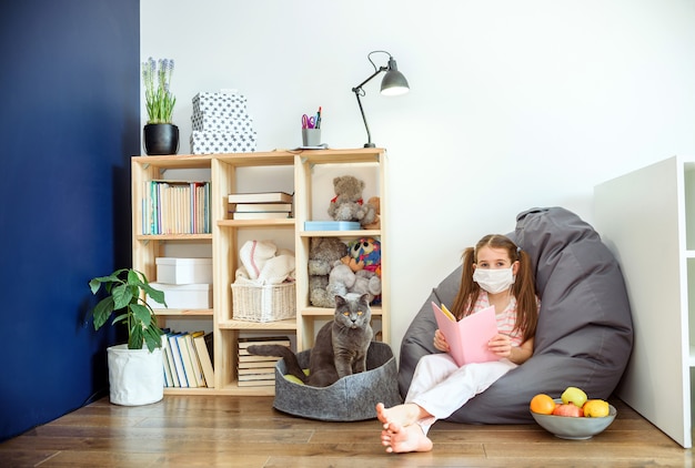 Foto una niña en máscara médica ubicando en el piso de madera y leyendo libros durante la cuarentena