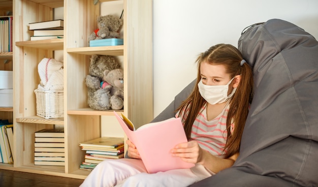 Una niña en máscara médica ubicando en el piso de madera y leyendo libros durante la cuarentena