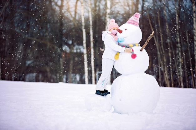 Una niña con una máscara y un muñeco de nieve jugando con un muñeco de nieve en un paseo invernal por la naturaleza