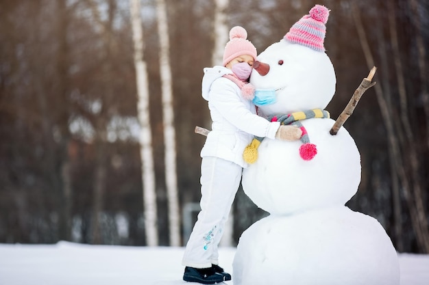 Una niña con una máscara y un muñeco de nieve jugando con un muñeco de nieve en un paseo invernal por la naturaleza