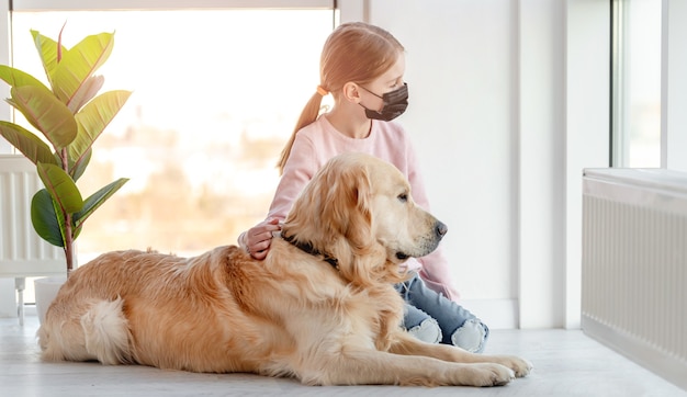 Niña con máscara negra y perro golden retriever sentados juntos en el suelo y mirando por la ventana