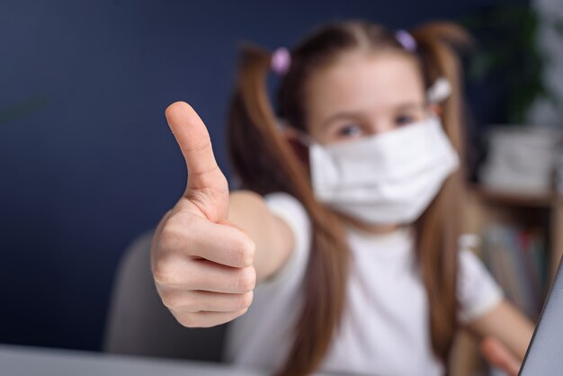 Una niña con máscara para proteger el coronavirus covid y mostrar los pulgares hacia arriba.