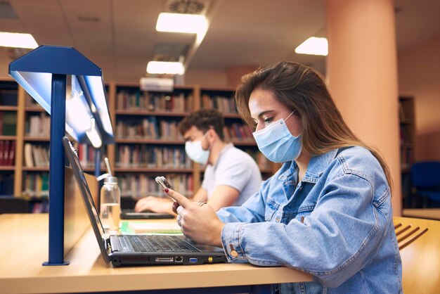 Foto niña con máscara usa un teléfono móvil mientras estudia en la biblioteca