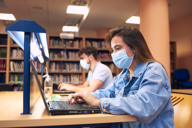 Una niña con máscaras estudia con computadoras portátiles en la biblioteca.