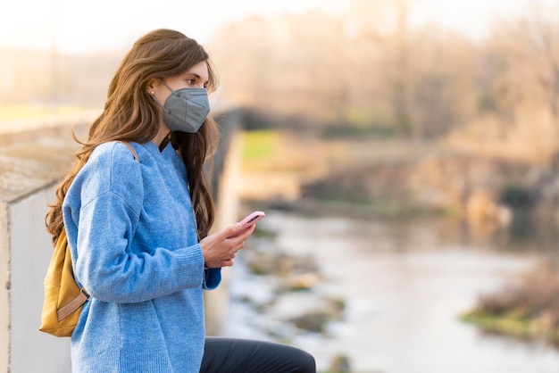 Niña con mascarilla gris mirando el teléfono mientras camina por el parque Lleva un suéter azul y una bolsa amarilla en la espalda Hay un río al fondo