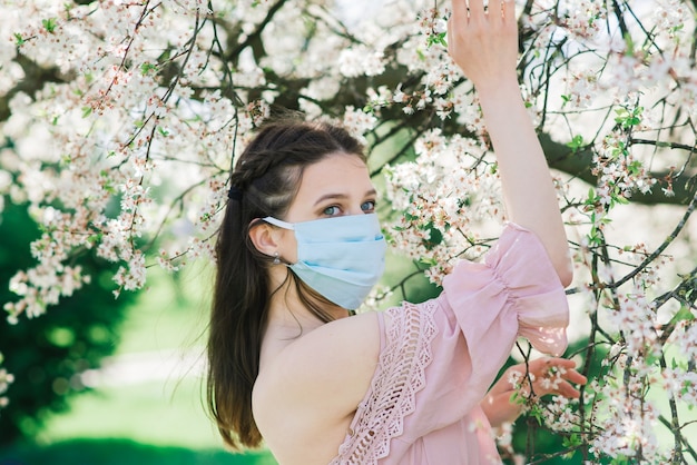 Una niña con una mascarilla médica para evitar la propagación del coronavirus en el parque entre árboles en flor.