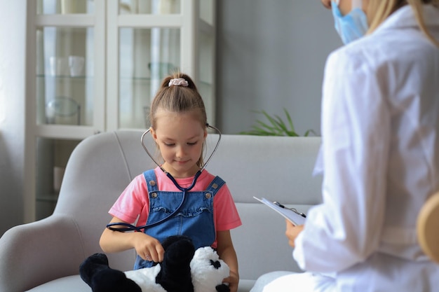 Niña en el médico para un chequeo. Doctor comprobando juguetonamente los latidos del corazón de un osito de peluche.