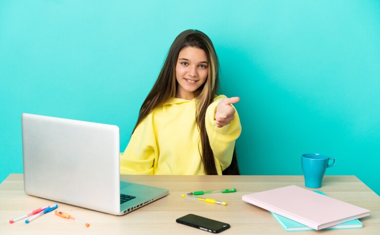 Foto niña en una mesa con un portátil sobre fondo azul aislado un apretón de manos para cerrar un buen trato