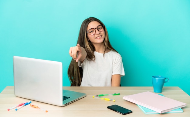 Niña en una mesa con un portátil sobre fondo azul aislado apuntando al frente con expresión feliz