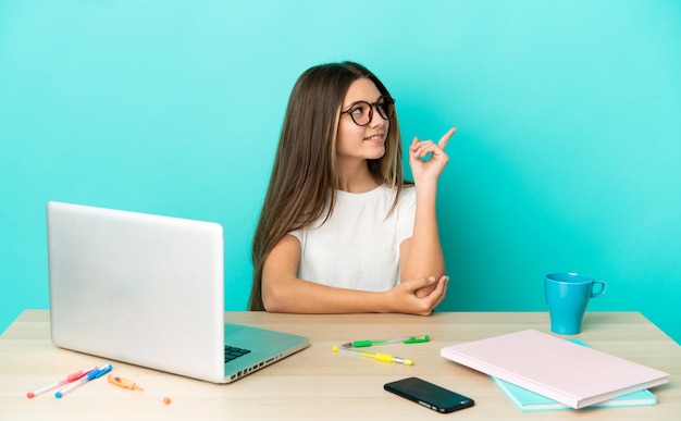 Niña en una mesa con un portátil sobre fondo azul aislado apuntando hacia una gran idea