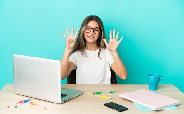 Niña en una mesa con un portátil sobre fondo azul aislado contando nueve con los dedos