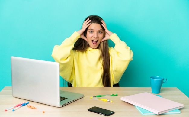 Niña en una mesa con un portátil sobre fondo azul aislado con expresión de sorpresa