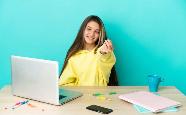 Niña en una mesa con un portátil sobre fondo azul aislado haciendo gesto de dinero